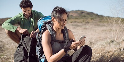 Dos excursionistas descansan en una zona seca y abierta; una persona sentada mira un teléfono móvil y lleva mochila de senderismo, mientras otra está detrás ajustando el equipo.
