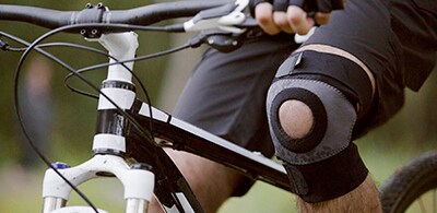 Persona montando una bicicleta con una rodillera negra de soporte en la rodilla derecha.
