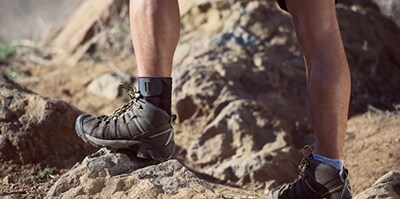Persona con botas de senderismo sobre rocas en un terreno montañoso, con una pierna apoyada en una piedra elevada y calcetines visibles.
